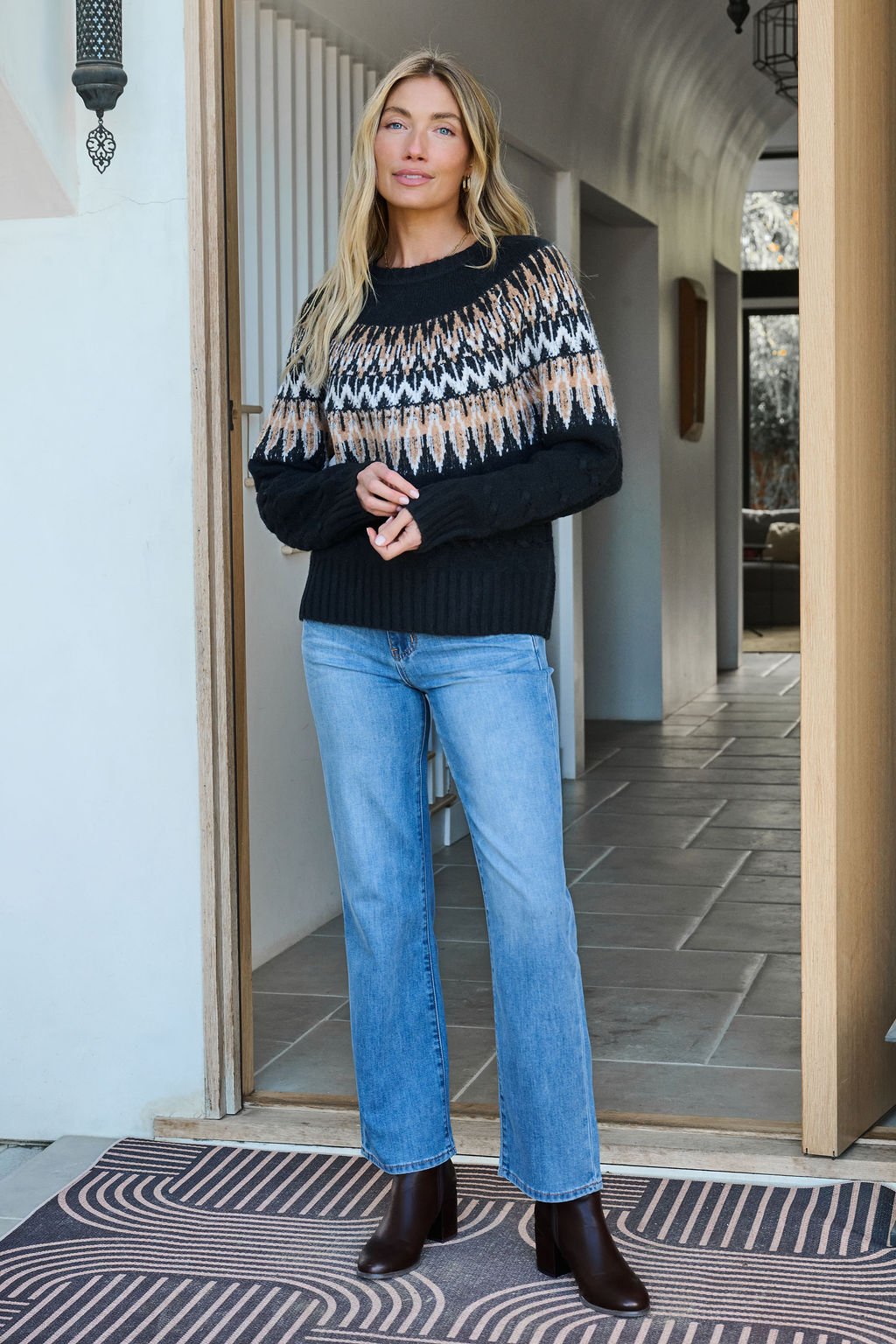 Woman wearing a Black Fair Isle Cable Knit Sweater stands in a doorway on a geometric rug, hands clasped.