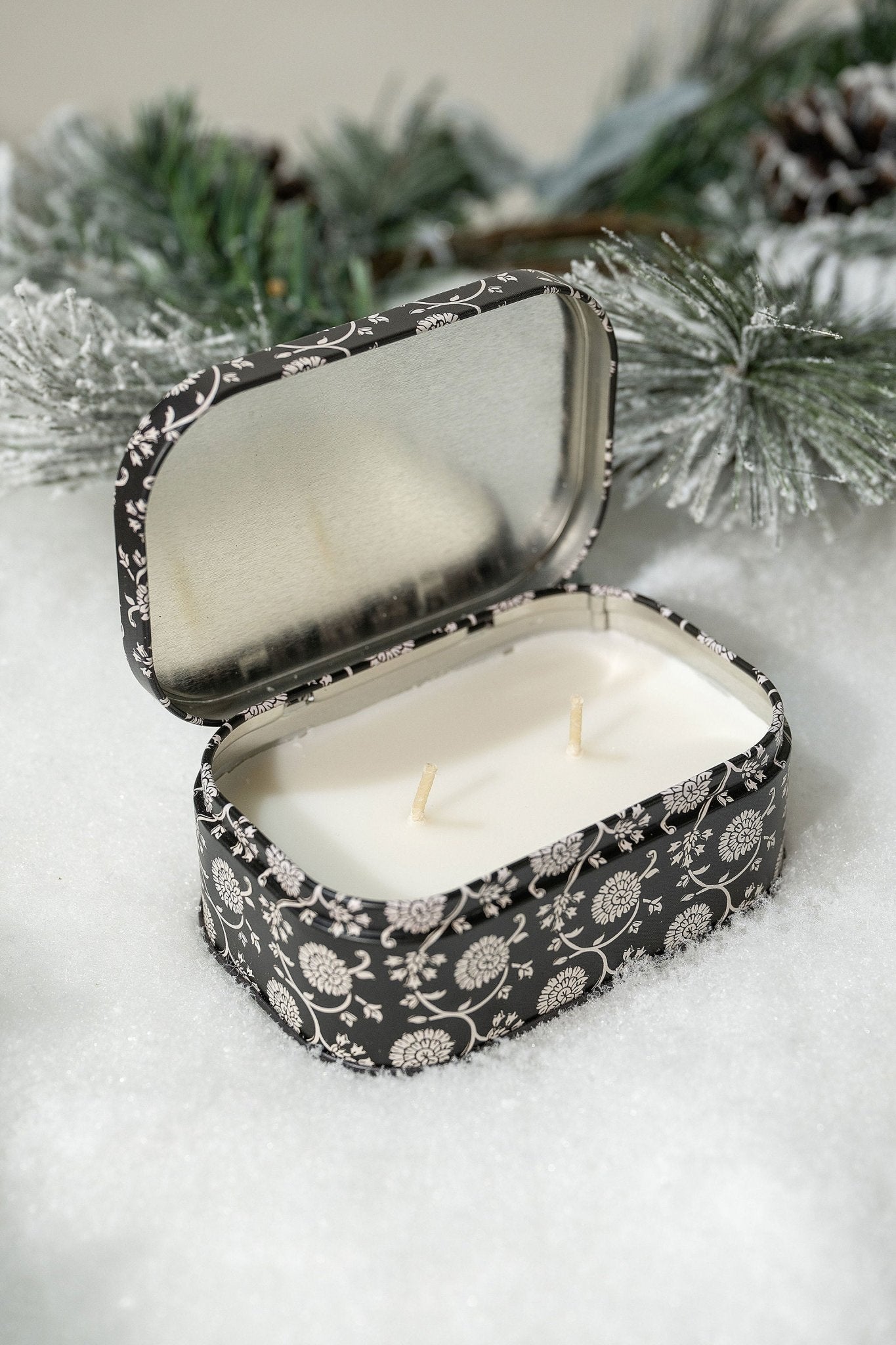 The Black Fig & Olive Print Tin Soy Candle—FINAL SALE—sits open on snow amid frosted pine branches.