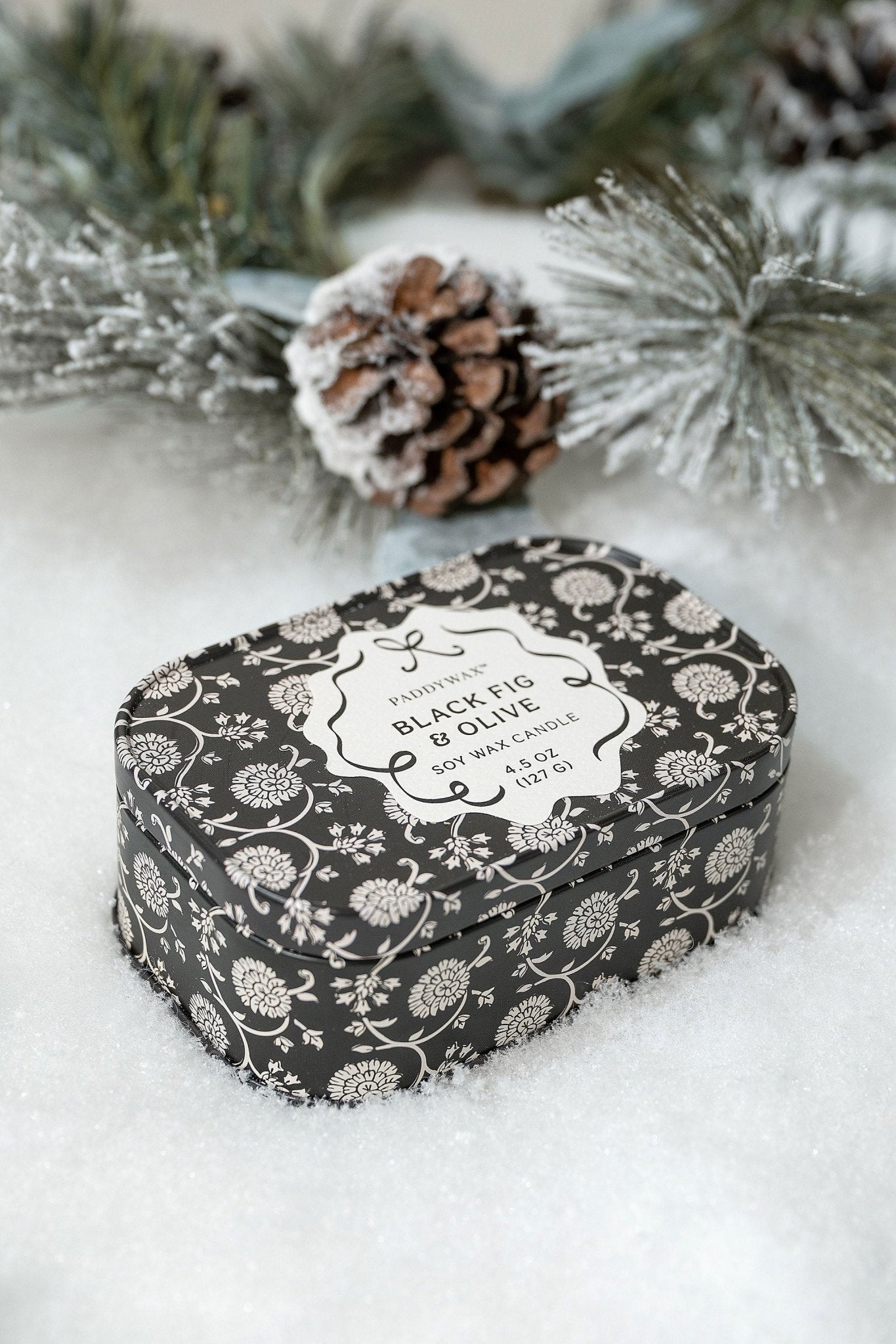 The Black Fig & Olive Print Tin Soy Candle - FINAL SALE rests on snow amid pine branches and a pinecone.