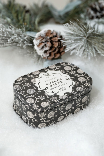 The Black Fig & Olive Print Tin Soy Candle - FINAL SALE rests on snow amid pine branches and a pinecone.