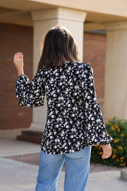 A person wearing the Black Floral Button Down Blouse - FINAL SALE and blue jeans stands outside near flowers, facing away.
