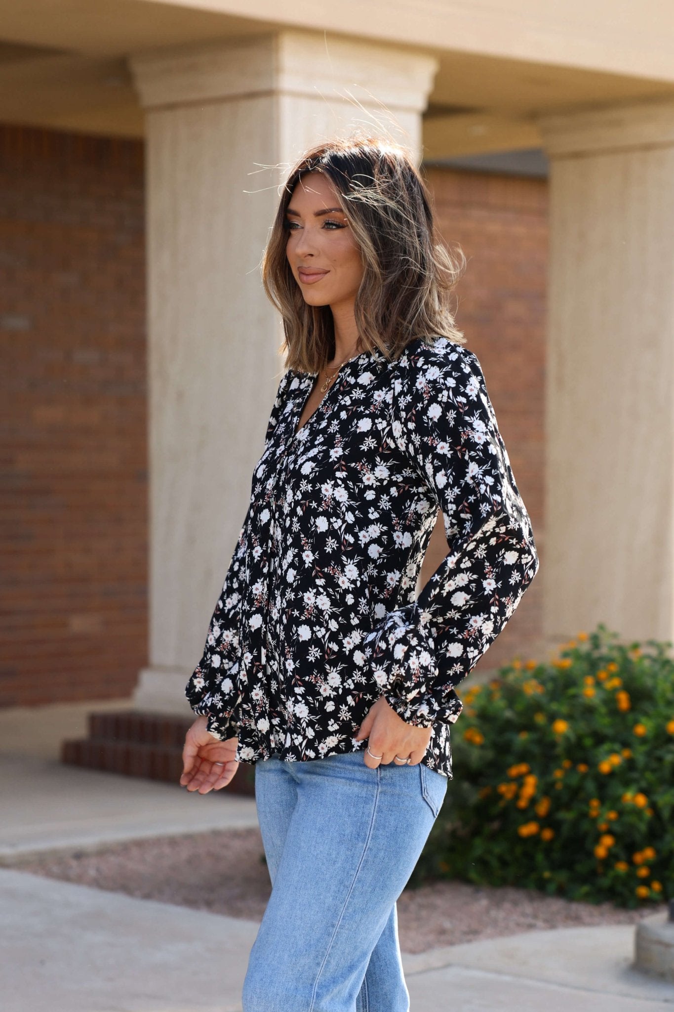 Woman with shoulder-length hair wears the Black Floral Button Down Blouse - FINAL SALE and light blue jeans outdoors near a building and flowers.