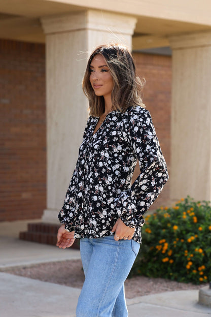Woman with shoulder-length hair wears the Black Floral Button Down Blouse - FINAL SALE and light blue jeans outdoors near a building and flowers.