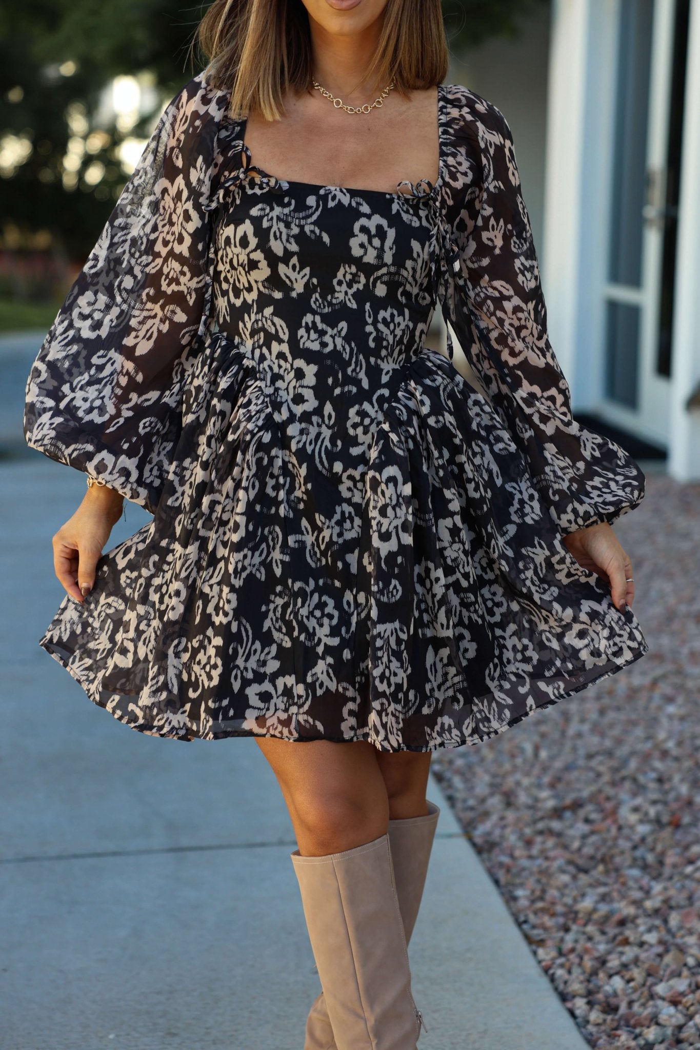 Woman wearing a Black Floral Chiffon Corset Mini Dress and tan knee-high boots, standing outside on a walkway.