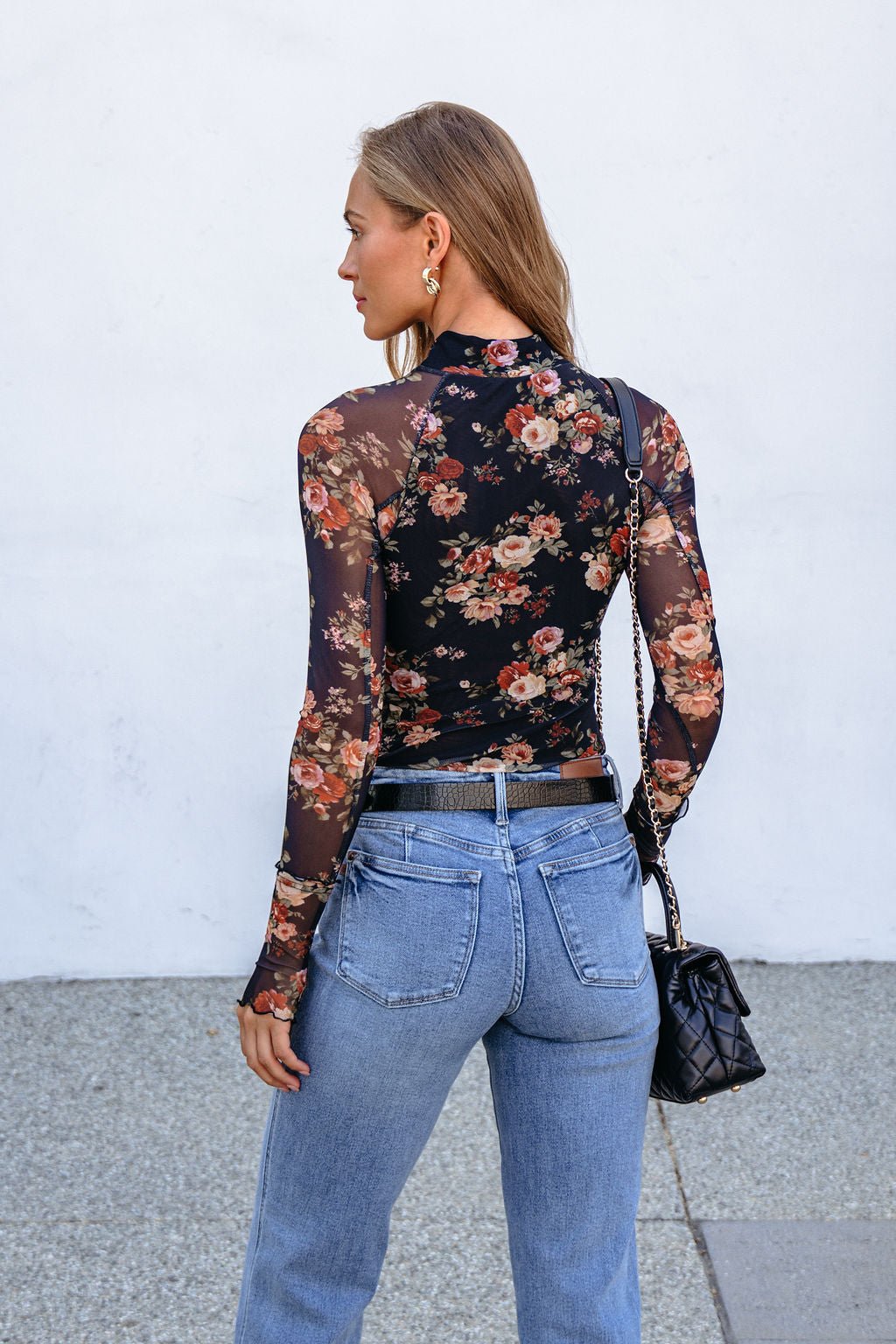 Woman facing away in the Black Floral Mesh Long Sleeve Top, light blue jeans, and a black quilted purse.