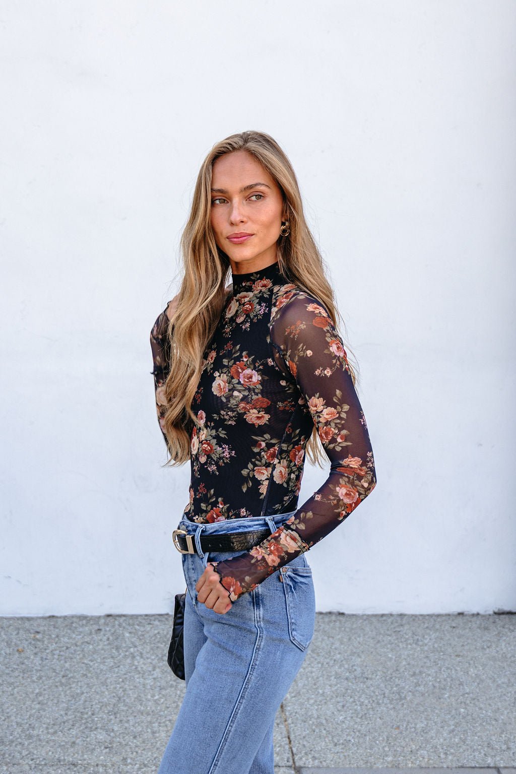 Woman in a Black Floral Mesh Long Sleeve Top with jeans stands outdoors, facing the camera against a white wall.