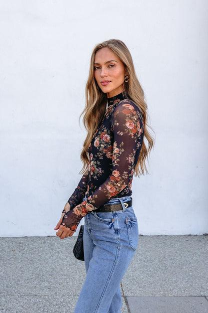 Woman wearing a Black Floral Mesh Long Sleeve Top and blue jeans stands outdoors, gazing at the camera.