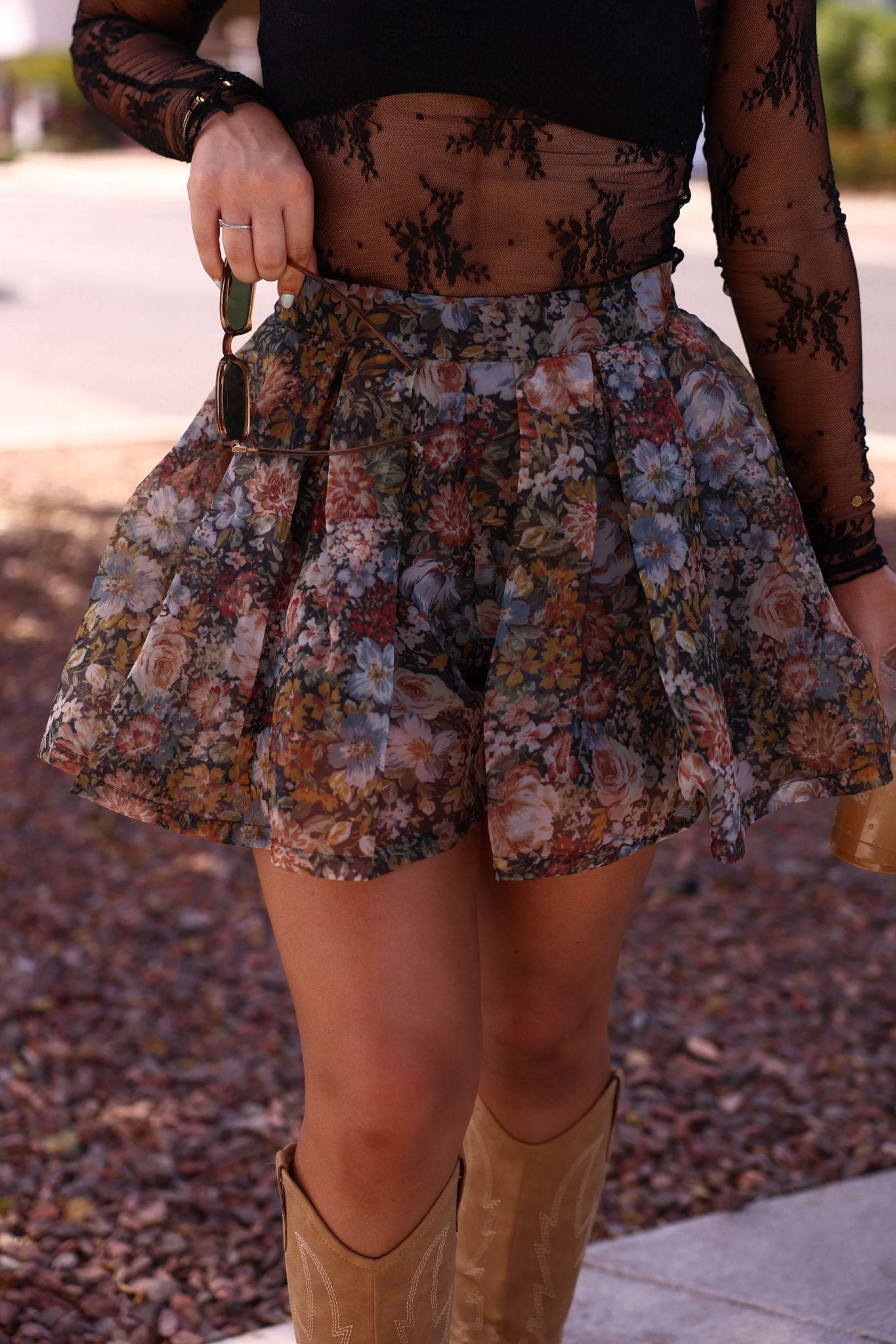 Person in a sheer black top and Black Floral Pleated Mini Skort, holding keys, with tan cowboy boots visible.