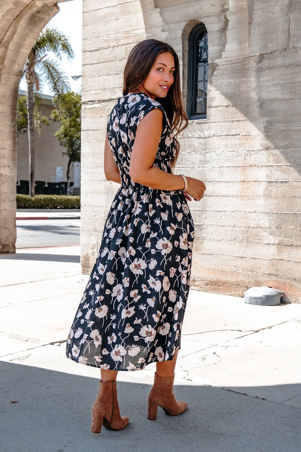 Woman in the Black Floral Print Smock Zip Midi Dress and brown heeled boots stands outdoors by a stone building, looking back.