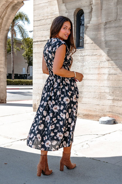 Woman in the Black Floral Print Smock Zip Midi Dress and brown heeled boots stands outdoors by a stone building, looking back.