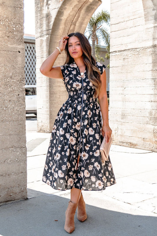 Woman wears a Black Floral Print Smock Zip Midi Dress and heels, outdoors by stone arches, holding a clutch.