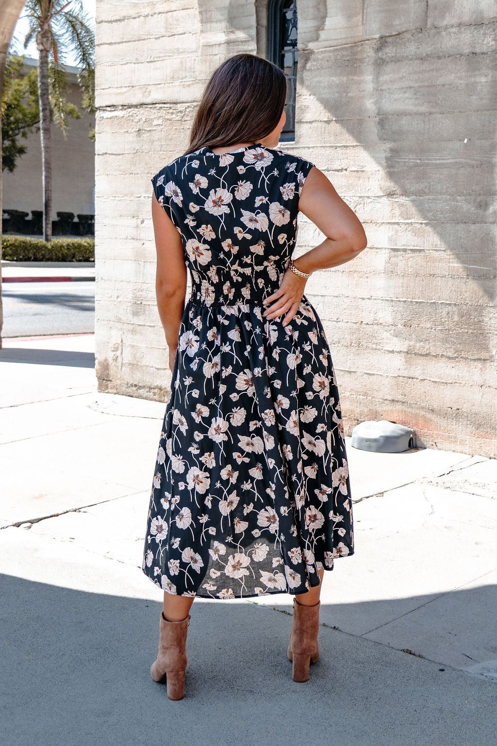 Woman wears a Black Floral Print Smock Zip Midi Dress with tan heels, standing outside by a sunlit concrete building.