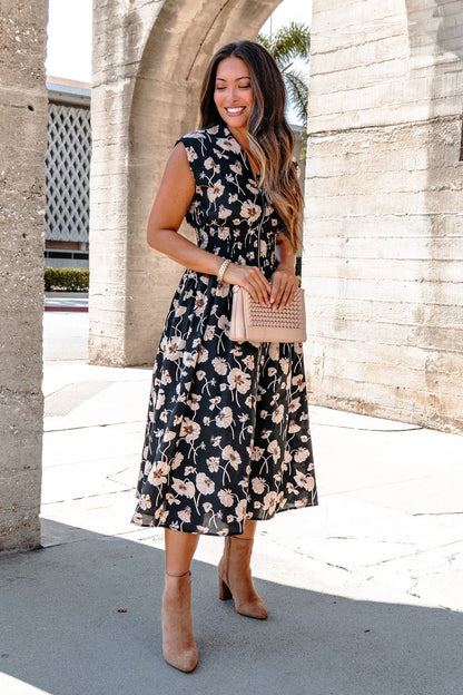 Woman in a Black Floral Print Smock Zip Midi Dress with tan heels stands outdoors, smiling and holding a light pink quilted purse.