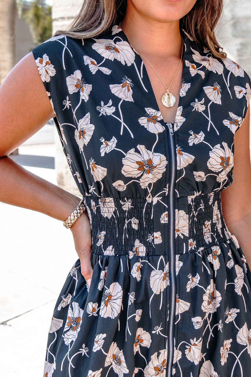 Woman wearing a Black Floral Print Smock Zip Midi Dress, gold necklace, and bracelet, standing outdoors.