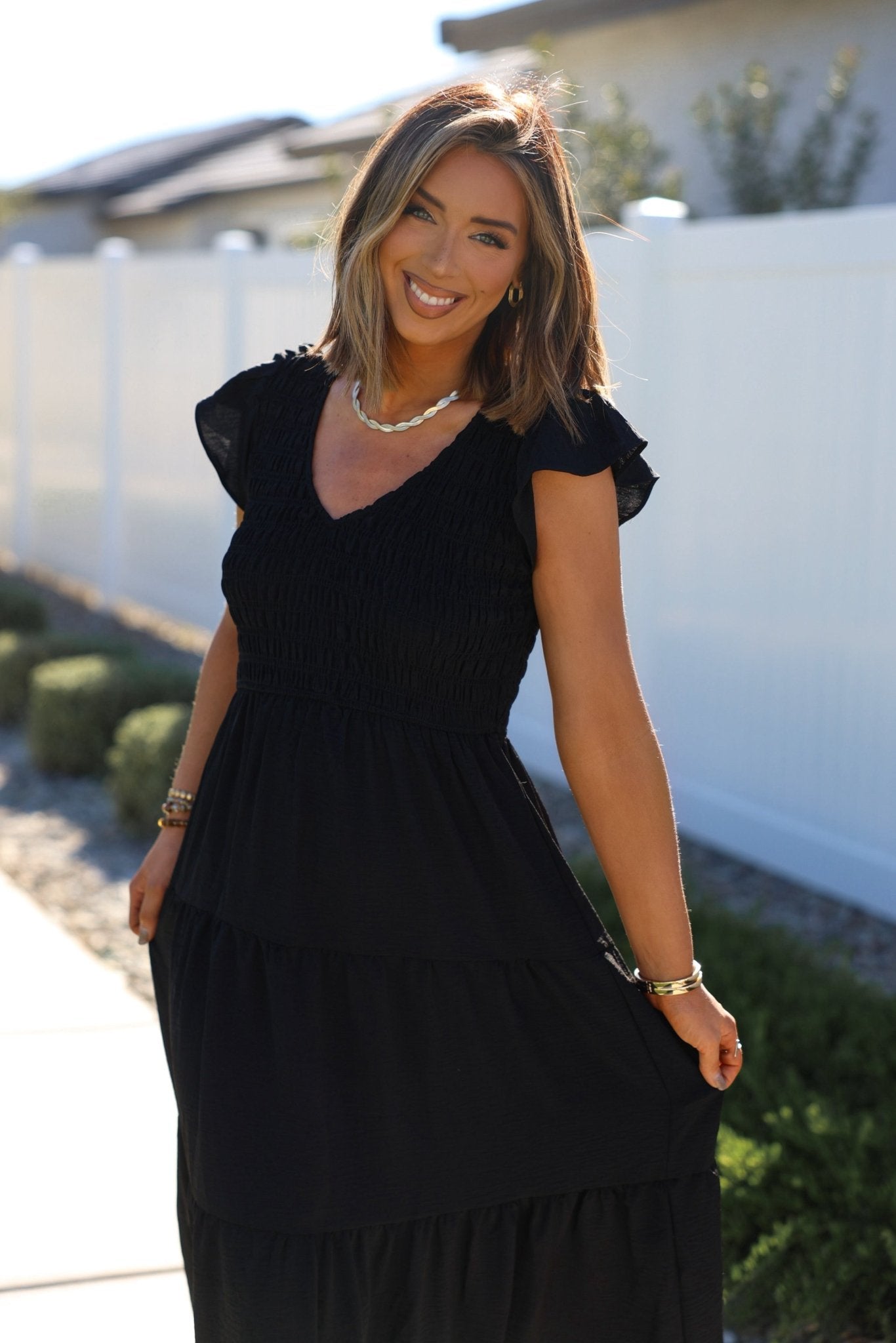 Woman smiling outdoors in a Black Flutter Sleeve Smock Tiered Midi Dress, standing by a white fence and greenery on a sunny day.