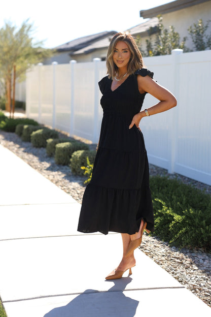 Woman in a Black Flutter Sleeve Smock Tiered Midi Dress and tan heels stands by a white fence, smiling at the camera.