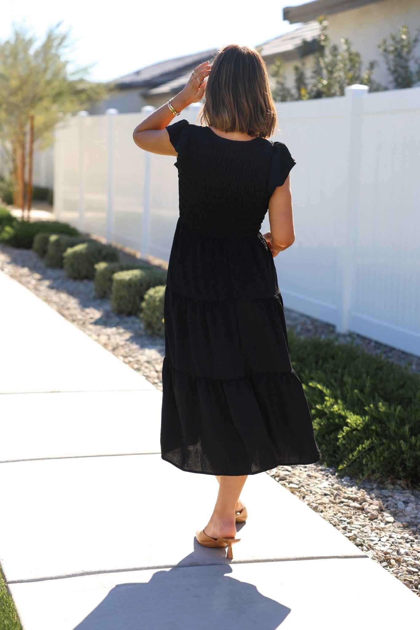 A woman wearing a Black Flutter Sleeve Smock Tiered Midi Dress walks on a sidewalk by a white fence and greenery, facing away.