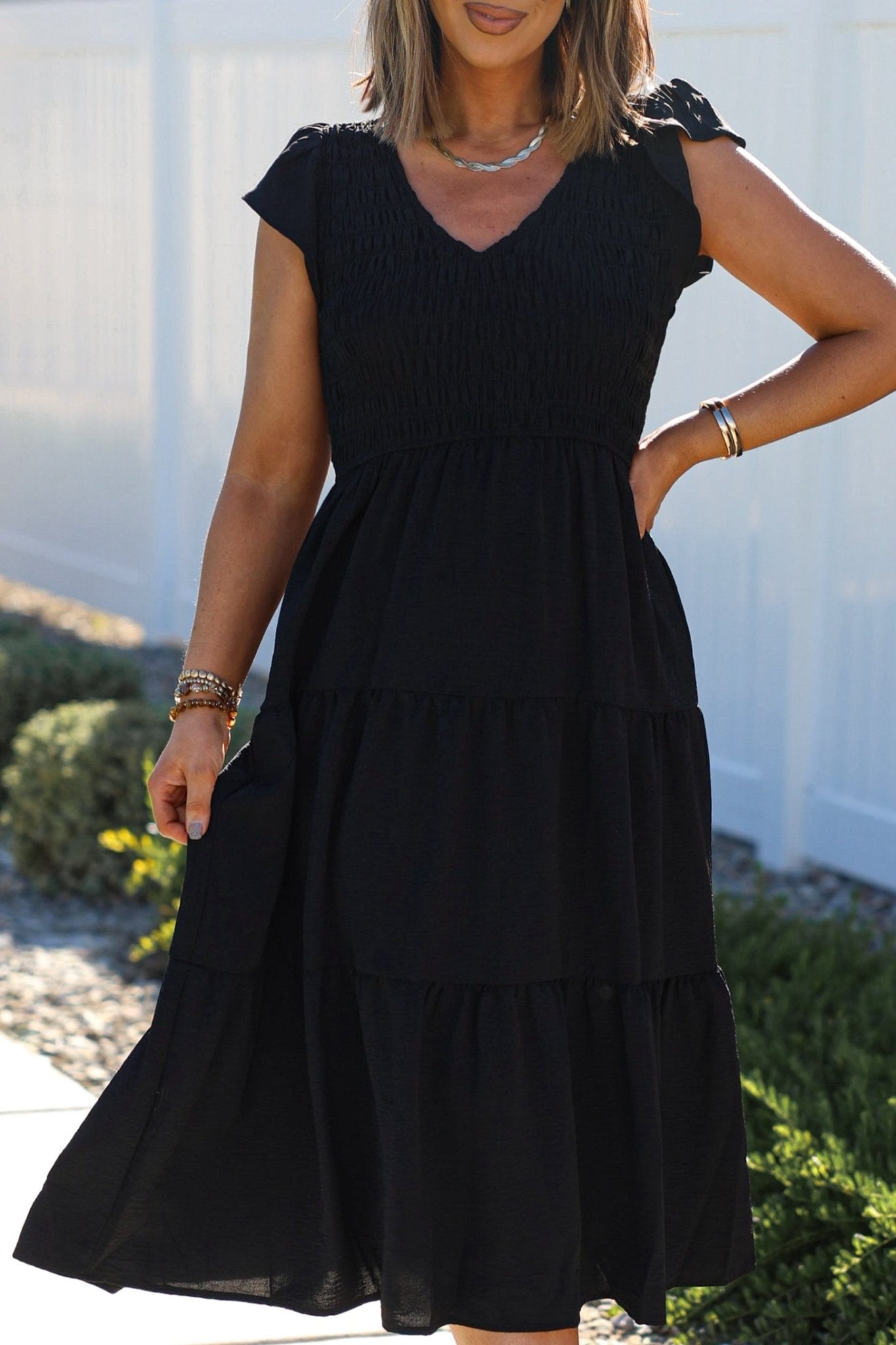 A woman stands outdoors by a white fence, wearing the Black Flutter Sleeve Smock Tiered Midi Dress.