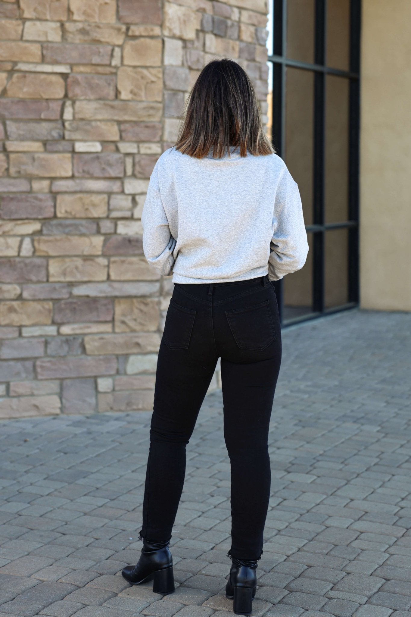 A woman in Black Frayed Hem Straight Leg Jeans and a gray sweatshirt stands outdoors facing a stone wall.