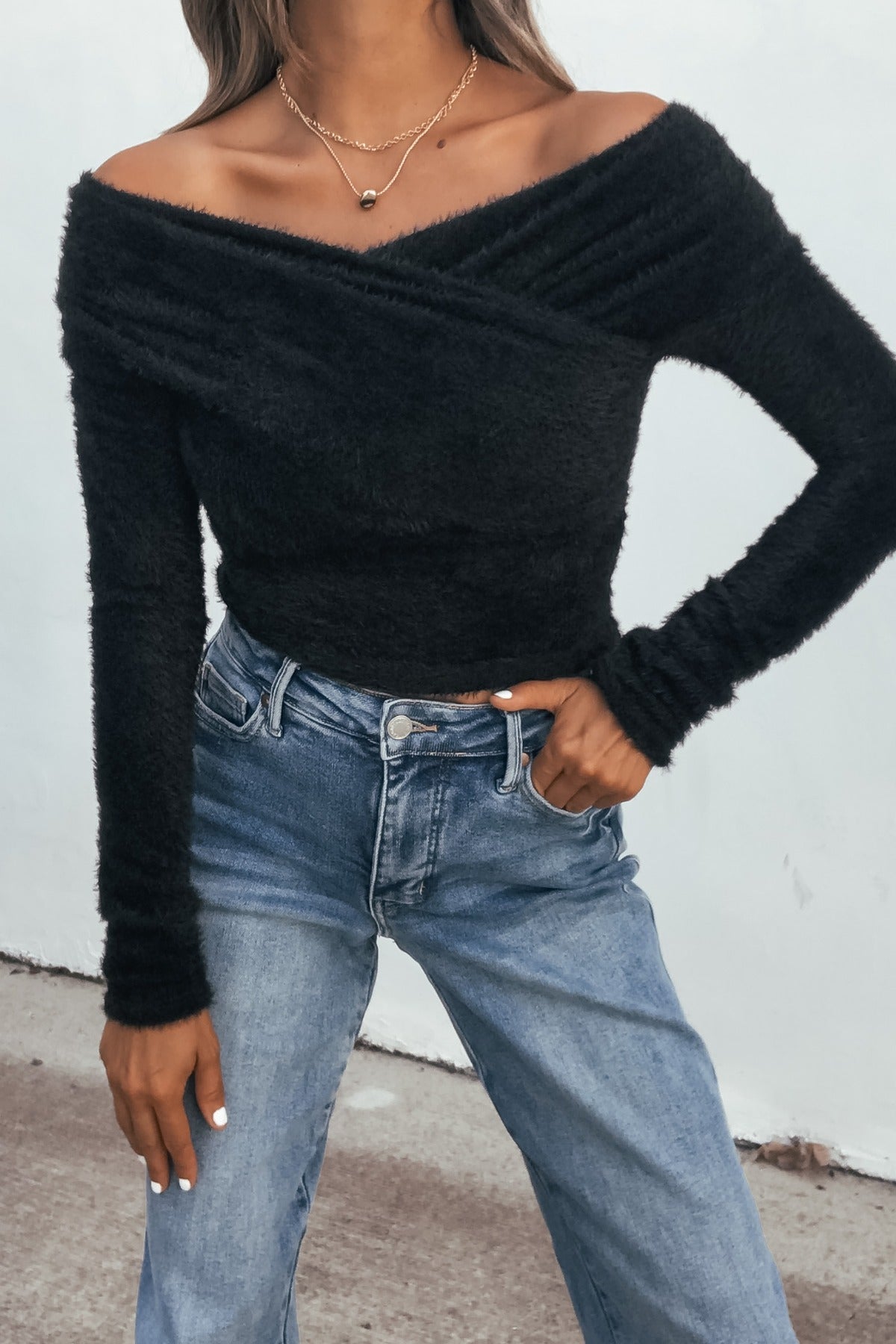 Woman in a black fuzzy knit off-shoulder top and light blue high-waisted jeans, standing outdoors.