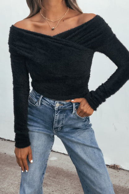 Woman in a black fuzzy knit off-shoulder top and light blue high-waisted jeans, standing outdoors.