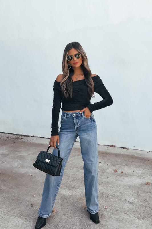 Woman in sunglasses wears a Black Fuzzy Knit Off-Shoulder Top, blue jeans, black heels, and holds a black quilted purse.