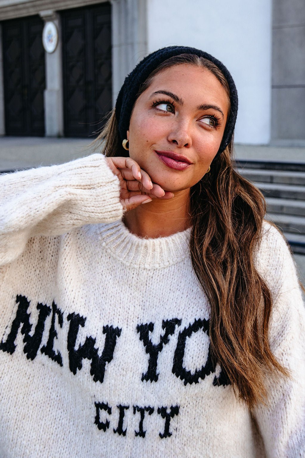Woman with long hair wears a white NYC sweater and the Black Fuzzy Soft Cable Knit Headband, looking up and smiling thoughtfully.