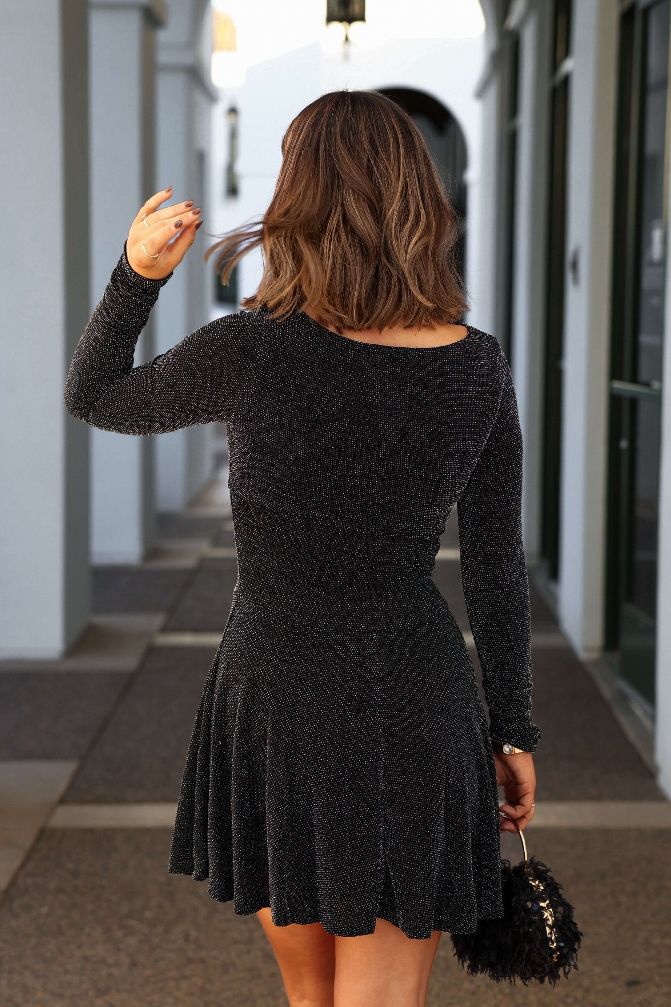 A woman outside, facing away, wears the Black Glittery Flare Hem Mini Dress-FINAL SALE, holding a small handbag and touching her hair.