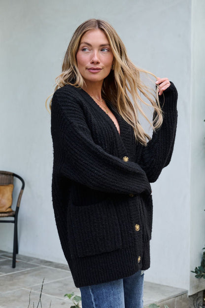 A woman in the Basic Black Knit Button Down Cardigan and jeans stands outside, looking to the side and touching her wavy blonde hair.