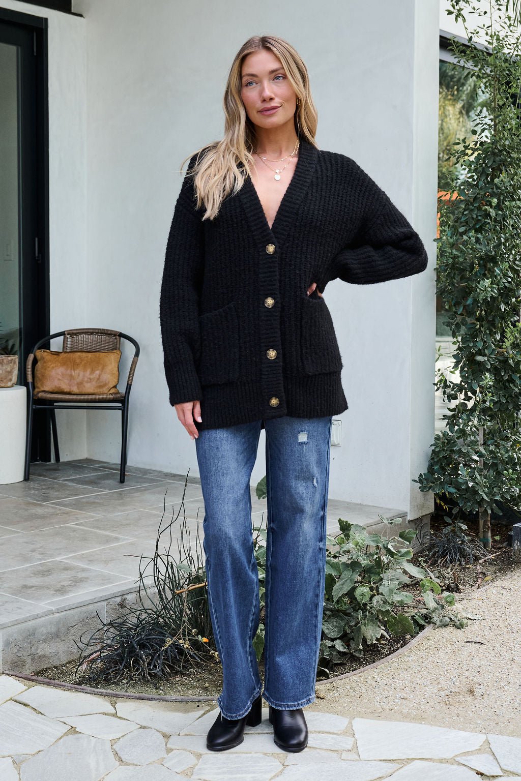 Woman outside a modern house in blue jeans and black boots, wearing our Basic Black Knit Button Down Cardigan for cozy winter style.