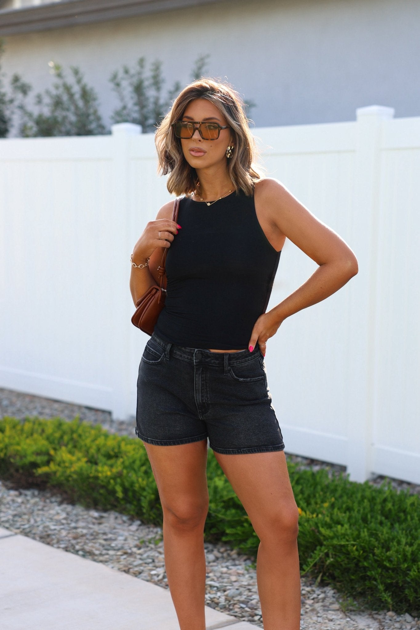 A woman in sunglasses and a black sleeveless top wears Black High Rise Classic Hem Shorts by a white fence with greenery.
