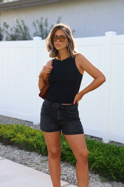 A woman in sunglasses and a black sleeveless top wears Black High Rise Classic Hem Shorts by a white fence with greenery.