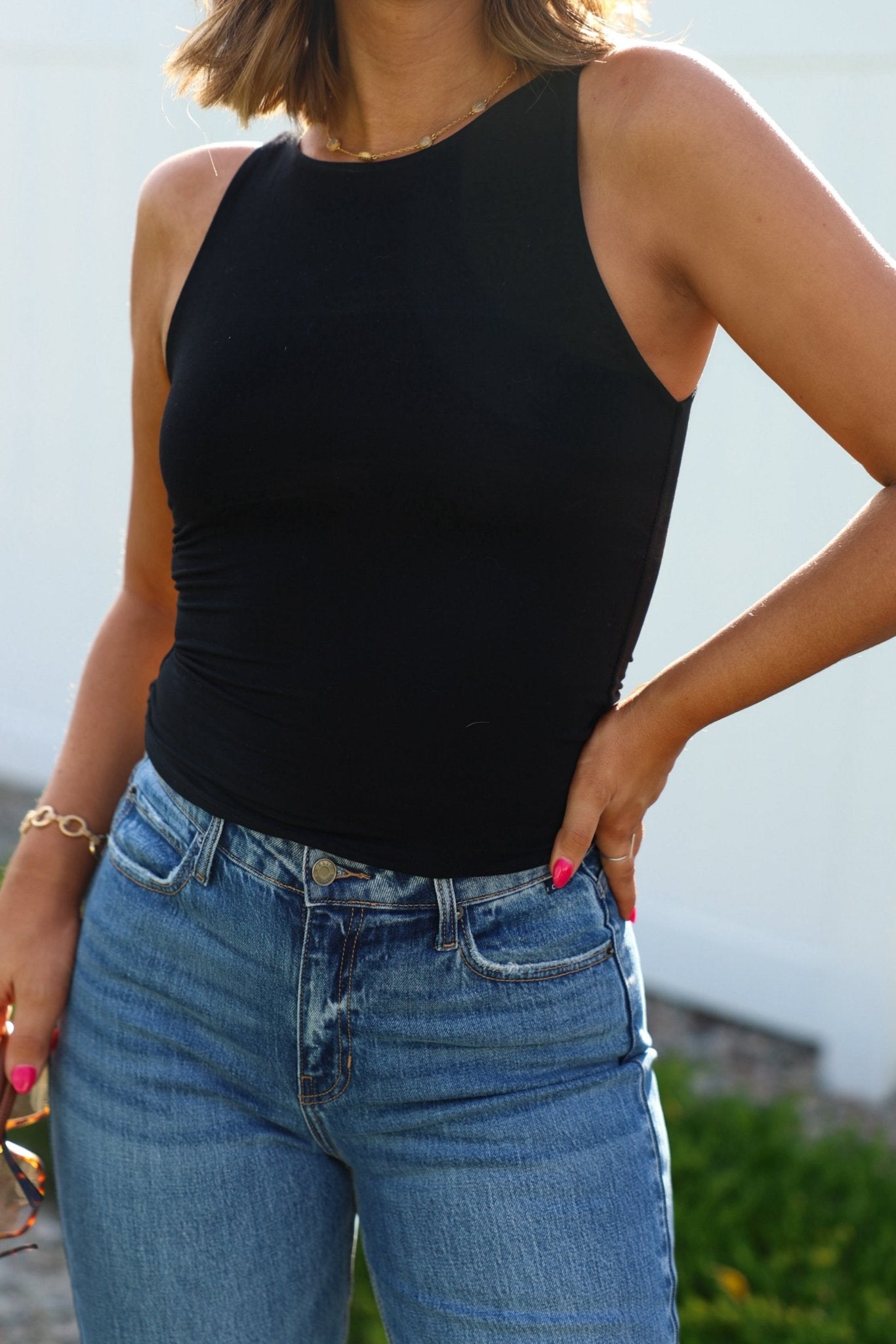 A woman wears a Black Jersey Knit Double Lined Tank Top and blue jeans, standing outside with one hand on her hip.
