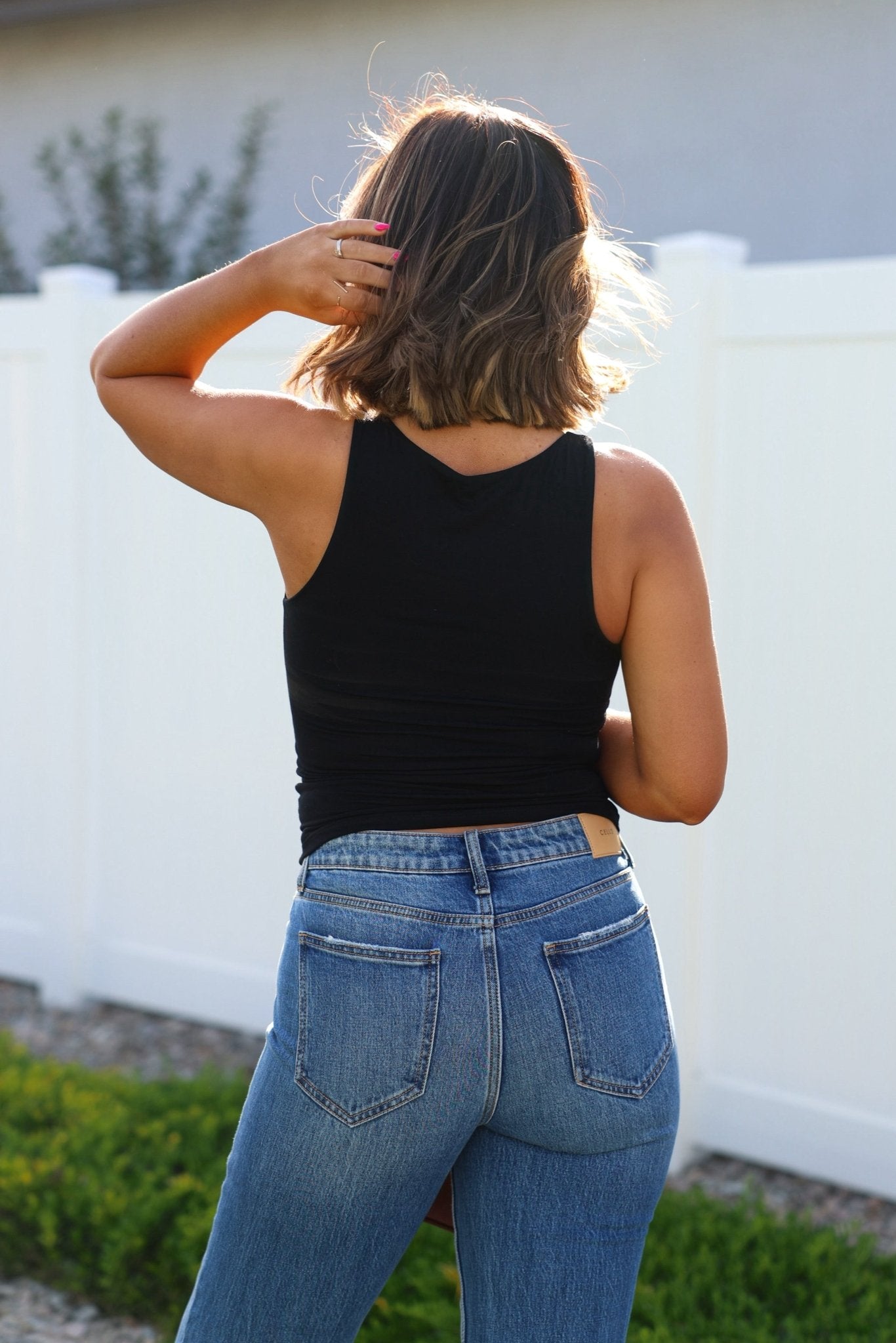 A woman with shoulder-length hair stands outside, facing away, in a Black Jersey Knit Double Lined Tank Top and blue jeans.