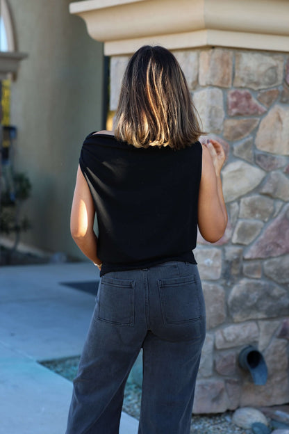A woman in a black jersey knit off-the-shoulder top and gray jeans stands outside, her shoulder-length hair framing her face.