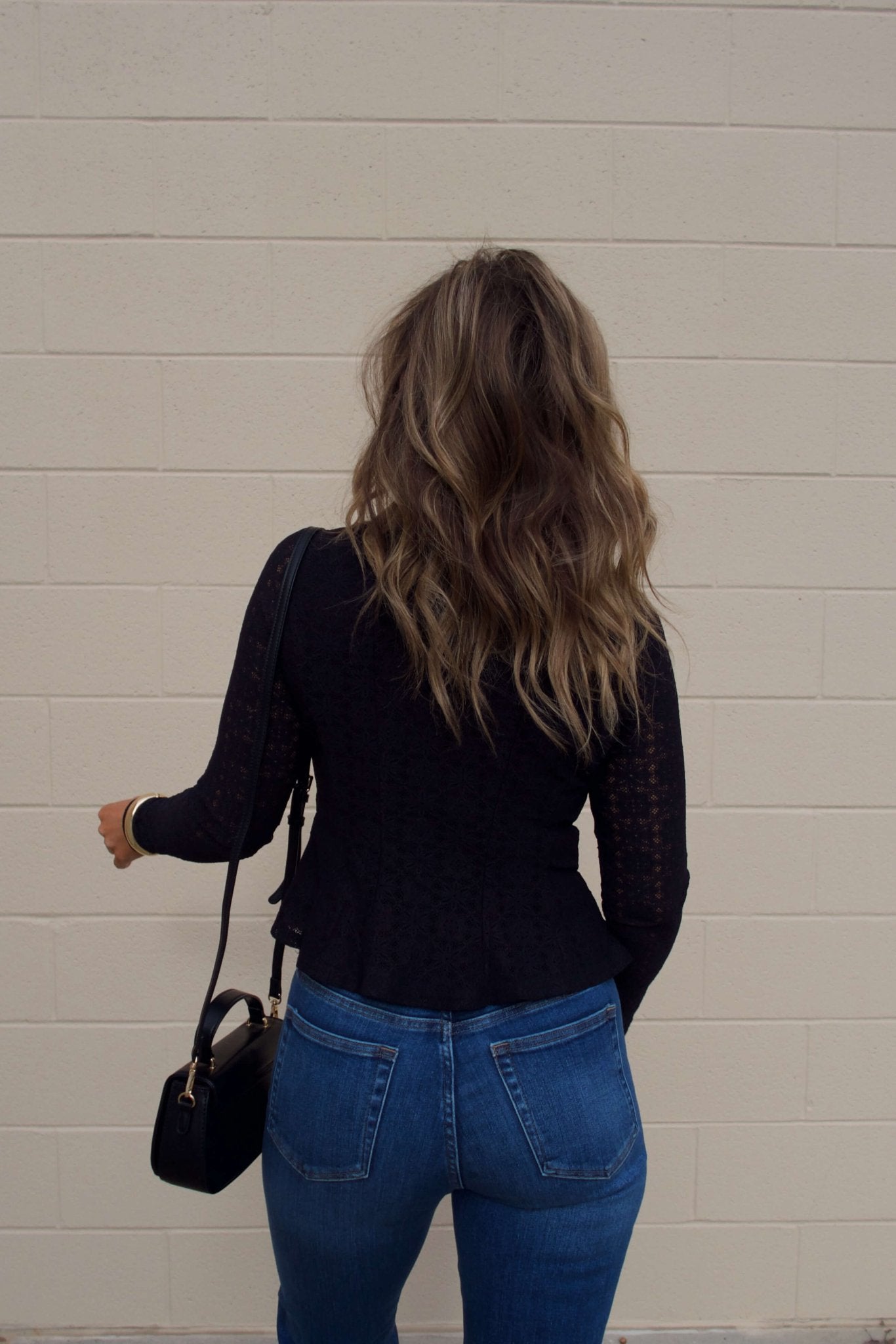 A person with wavy brown hair in the Black Lace Princess Seam Top and blue jeans stands by a beige brick wall, holding a black purse.