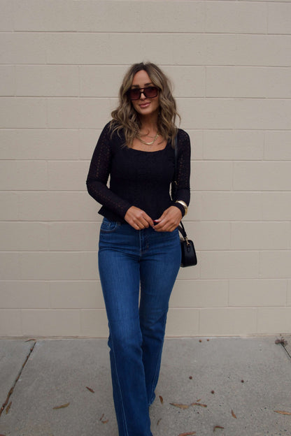 A woman with wavy hair wears the Black Lace Princess Seam Top and stands in front of a beige brick wall.