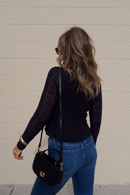 A woman with wavy hair wears the Black Lace Princess Seam Top, blue jeans, sunglasses, and carries a black purse by a beige brick wall.