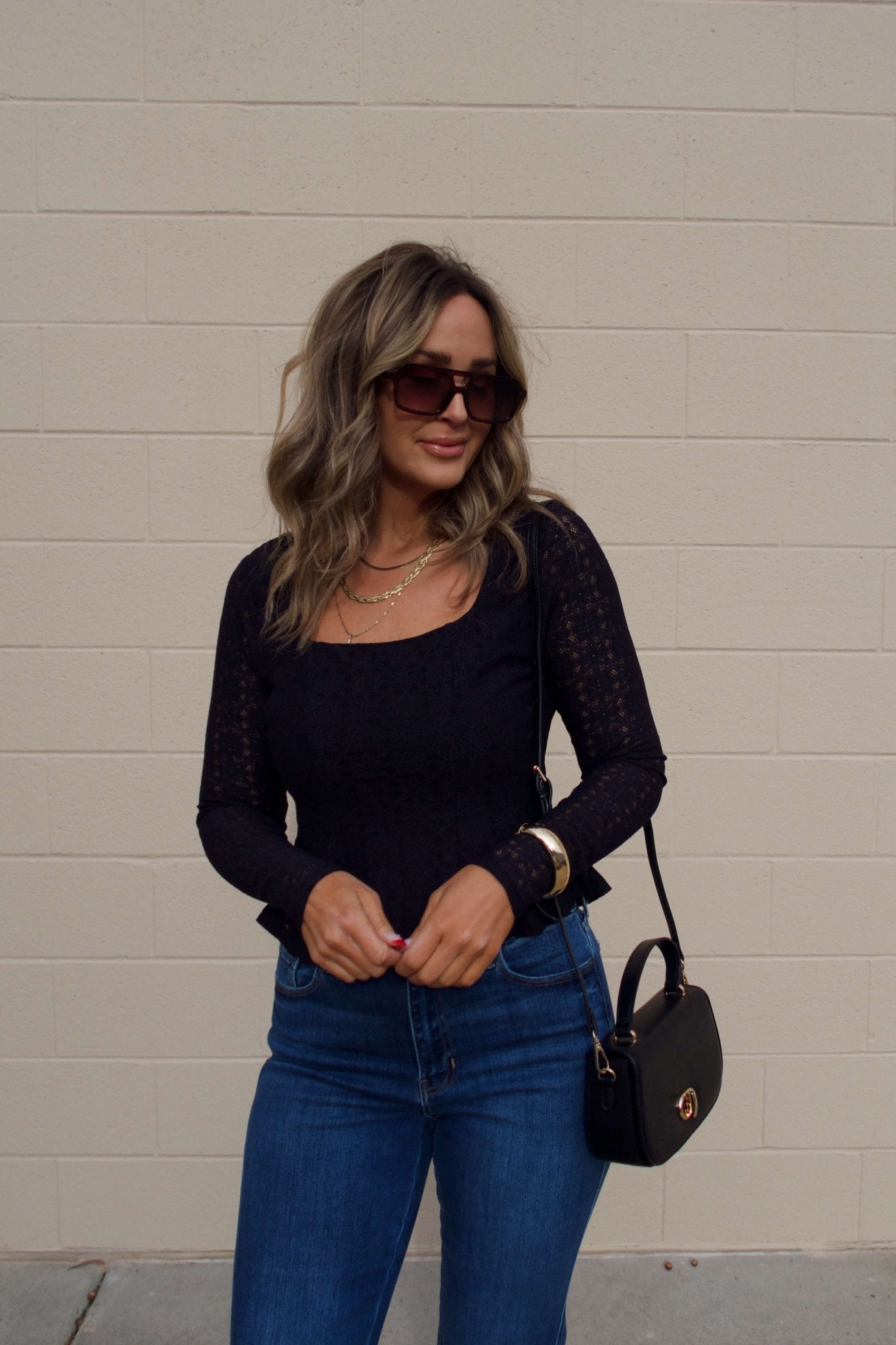 Woman with wavy hair wearing the Black Lace Princess Seam Top stands against a beige brick wall, holding a small black purse.