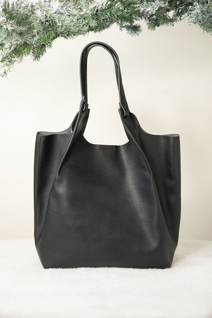 The Black Maeve Pebbled Leather Tote Bag rests on a white surface, accented by frosted evergreen branches above.