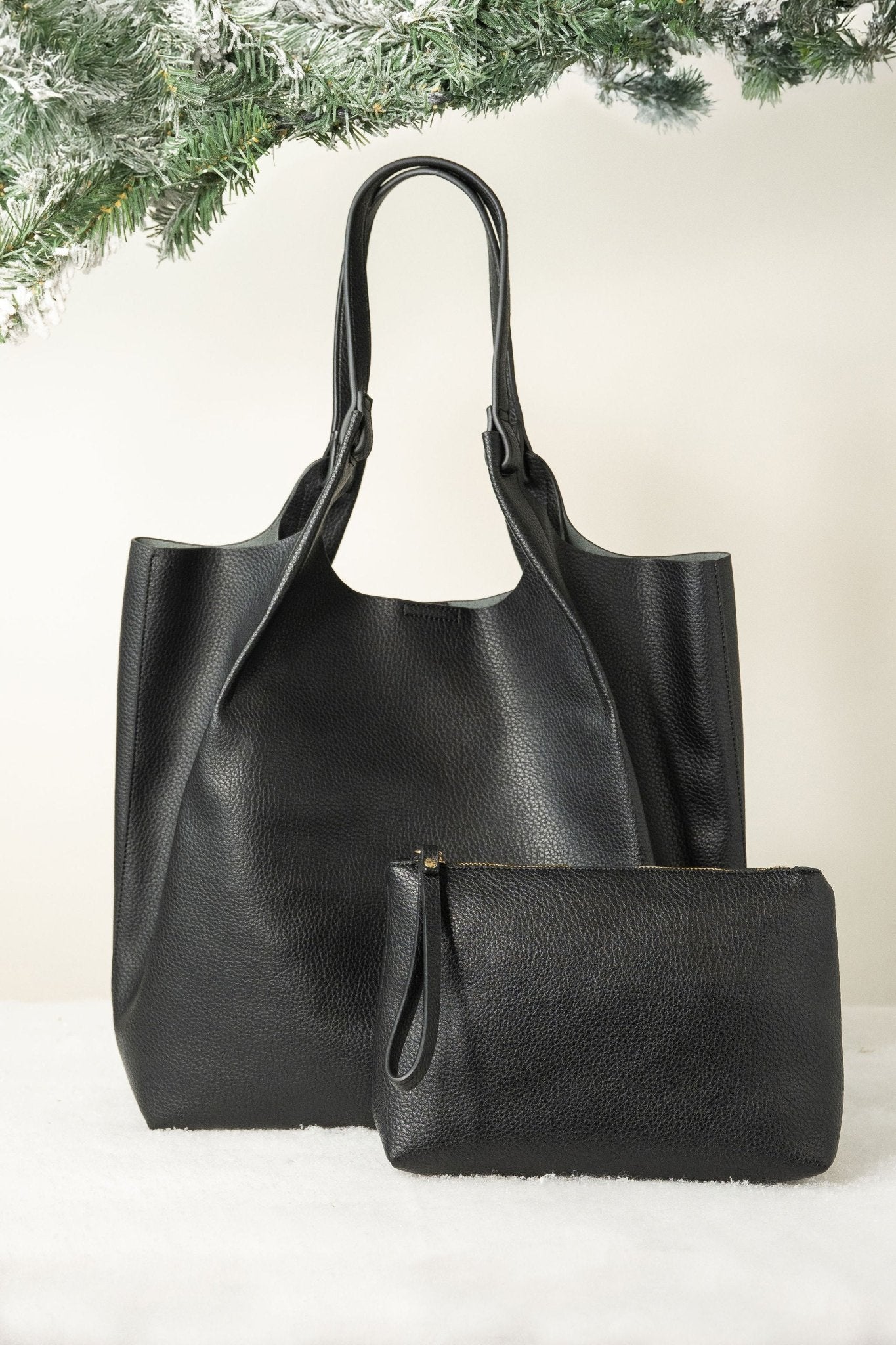 The Black Maeve Pebbled Leather Tote Bag is displayed with a matching pouch on white, accented by pine branches for a chic look.