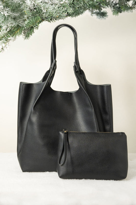 The Black Maeve Pebbled Leather Tote Bag is displayed with a matching pouch on white, accented by pine branches for a chic look.