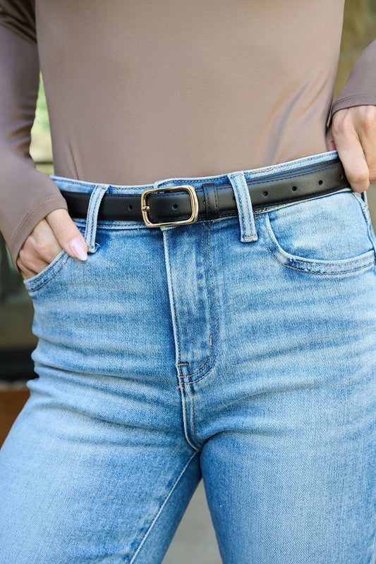 Person wearing light blue jeans, taupe long-sleeve top, and Black Metallic Buckle Leather Belt, hands in front pockets.