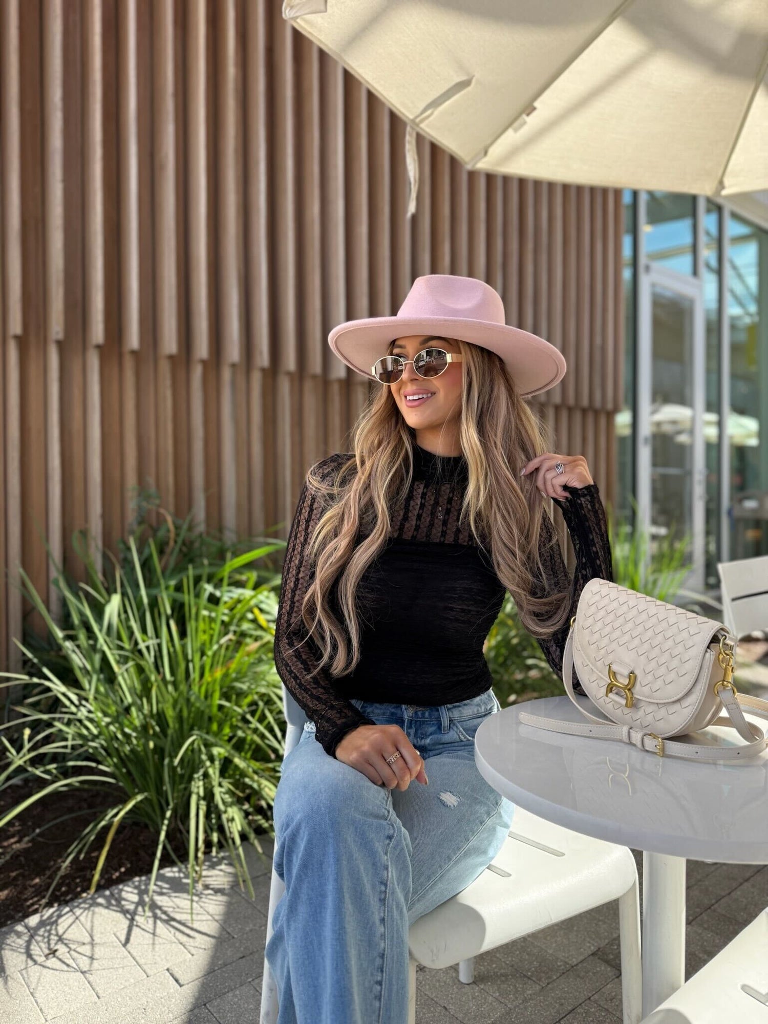 A woman in sunglasses and jeans smiles at an outdoor café in the Black Mock Neck Sheer Lace Bodysuit, holding a white handbag.