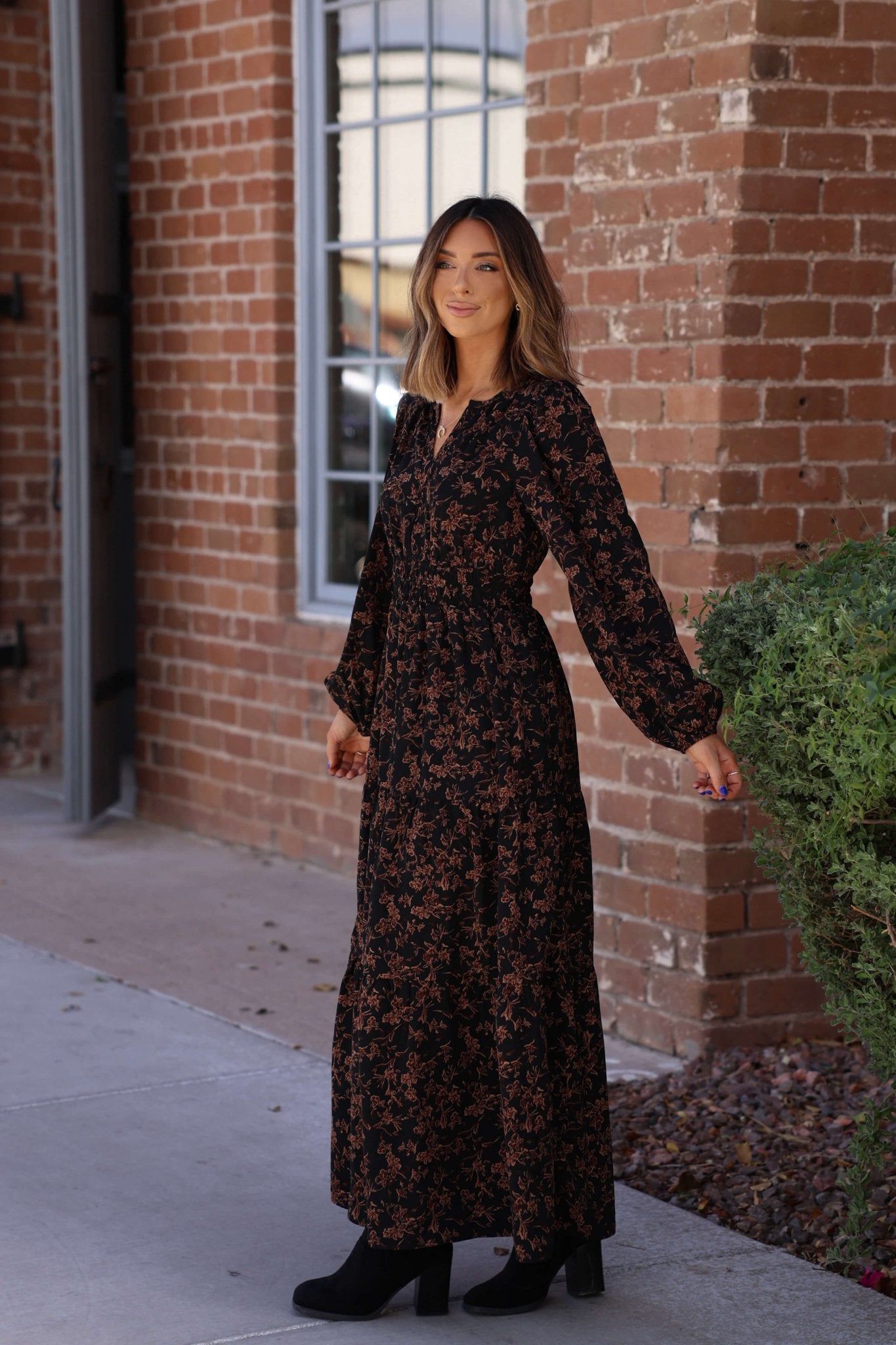A woman wears the Black Multi Floral Print Tiered Midi Dress outside a brick building, smiling and holding the skirt.