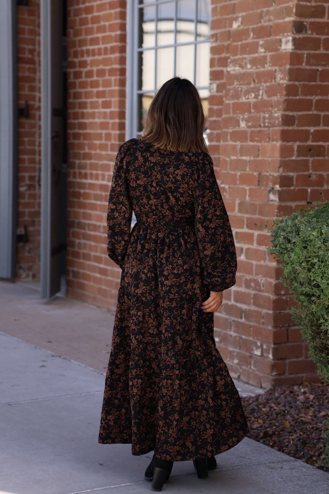 A woman wears a Black Multi Floral Print Tiered Midi Dress and boots, standing outside near a brick building and green shrub.
