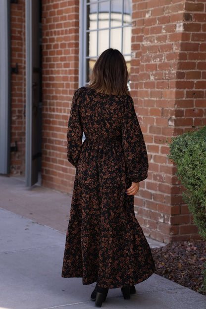 A woman wears a Black Multi Floral Print Tiered Midi Dress and boots, standing outside near a brick building and green shrub.