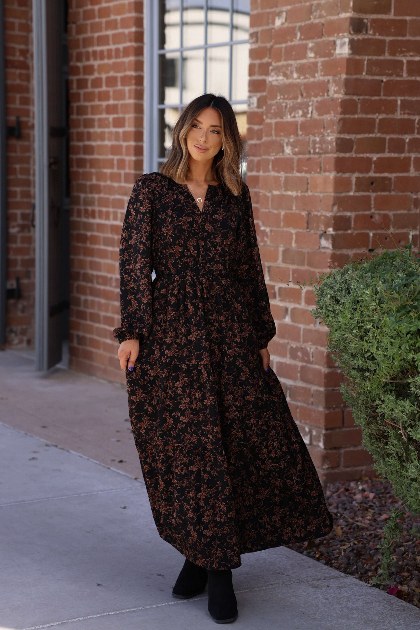 A woman stands outside by a brick wall in the Black Multi Floral Print Tiered Midi Dress.