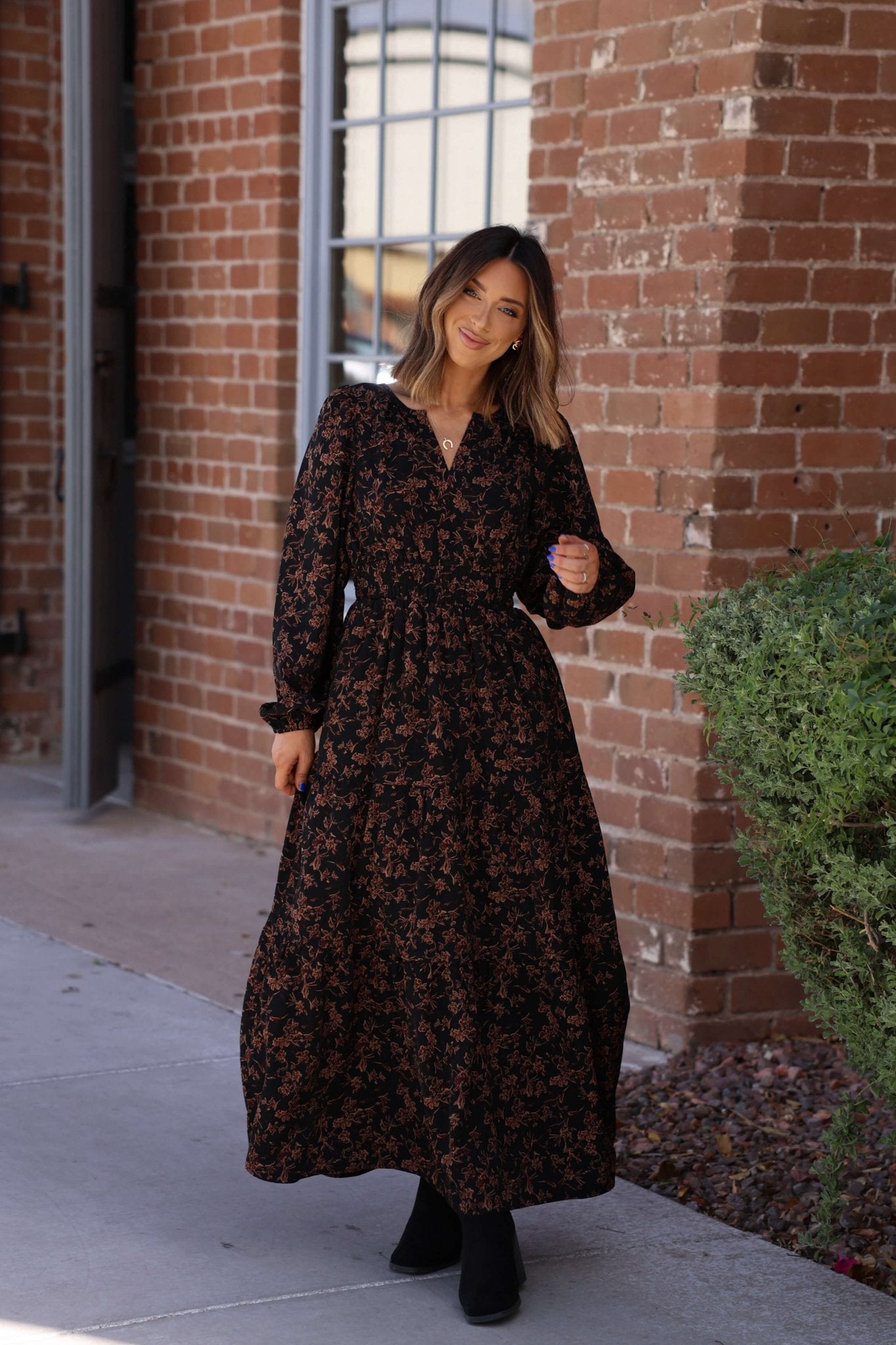 A woman wearing a Black Multi Floral Print Tiered Midi Dress smiles on a sidewalk near a brick building and green bushes.