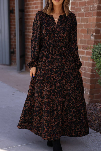 Woman in a Black Multi Floral Print Tiered Midi Dress with long sleeves, standing on a sidewalk by a brick wall.
